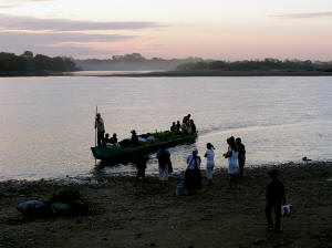 Long canoe-like boat landing at dark riverbank in front of distant sunset